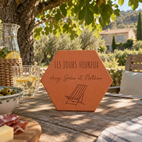 Tomette personnalisée « Les jours heureux chez Julie et Nathan » et chaise longue gravées, sur une table de jardin près de verres et d’olives.