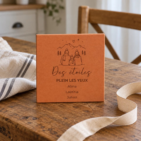 Carreau personnalisé « Des étoiles plein les yeux », famille en montagne et trois prénoms gravés, dressé sur une table en bois près d’un ruban et d’un torchon.