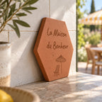 Tomette gravée « La Maison du Bonheur », parasol et chaise longue, vue de côté sur une table de terrasse, près d’une branche d’olivier.