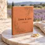 Carreau de mariage personnalisé Emma et Jules, date et feuillages gravés, vu de biais sur un support rond près des alliances et d’un tissu clair.