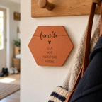 Tomette personnalisée « famille », cœur et quatre prénoms gravés, fixée au mur sous un porte-manteau, près d’un sac.