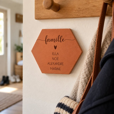 Tomette personnalisée « famille », cœur et quatre prénoms gravés, fixée au mur sous un porte-manteau, près d’un sac.