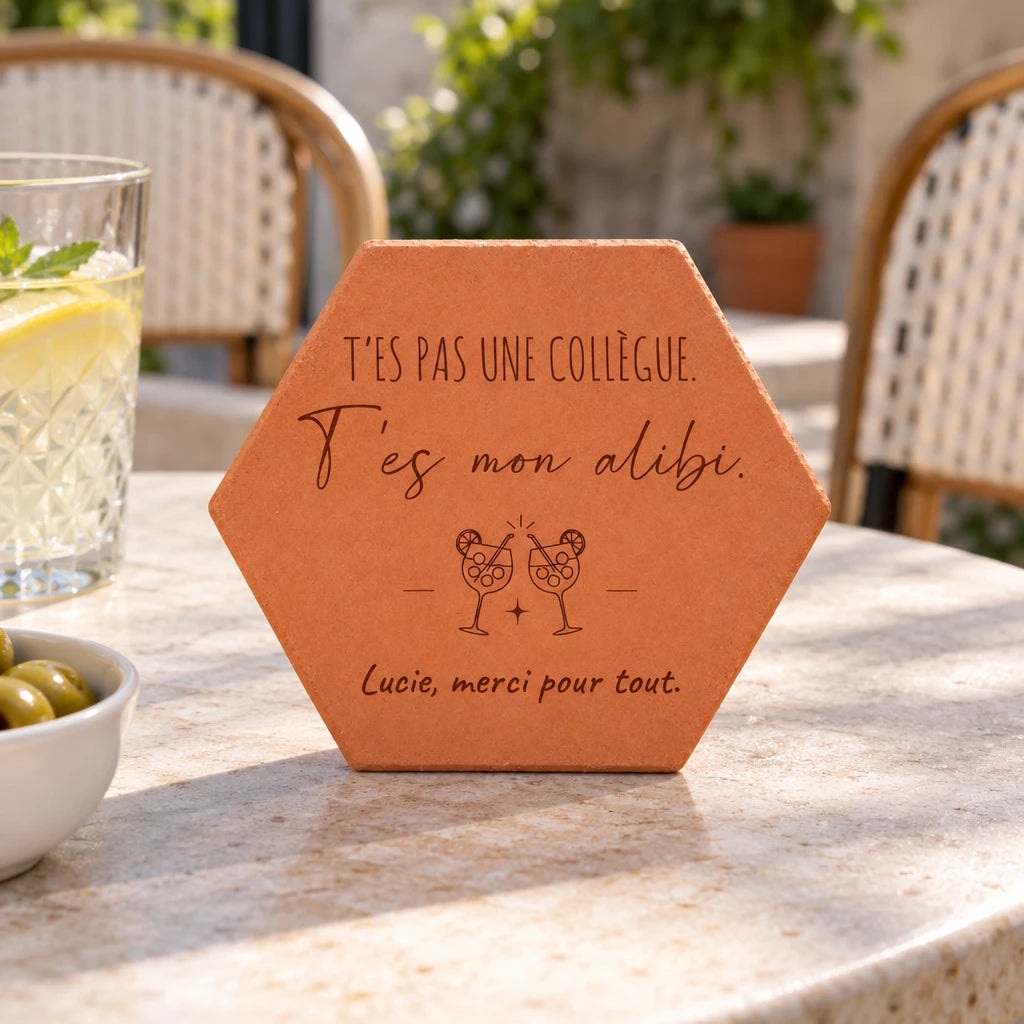 Pieza hexagonal de terracota «T'es mon alibi» sobre una mesa de terraza junto a un vaso con limón y un cuenco de aceitunas.