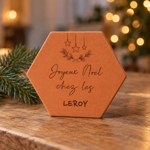 Tomette « Joyeux Noël chez les Leroy » posée sur une table en bois, devant un sapin illuminé et des branches de sapin.