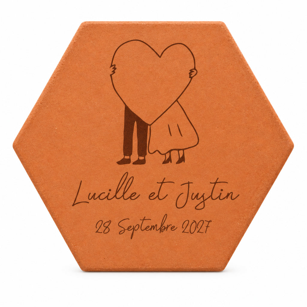 Pieza hexagonal de terracota grabada con una pareja escondida tras un gran corazón, los nombres Lucille y Justin y una fecha, sobre fondo blanco.