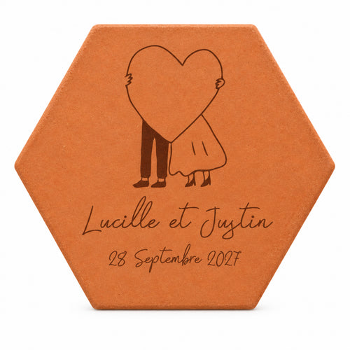 Hexagonal terracotta tile engraved with a couple hiding behind a big heart, the names Lucille and Justin and a date, on a white background.