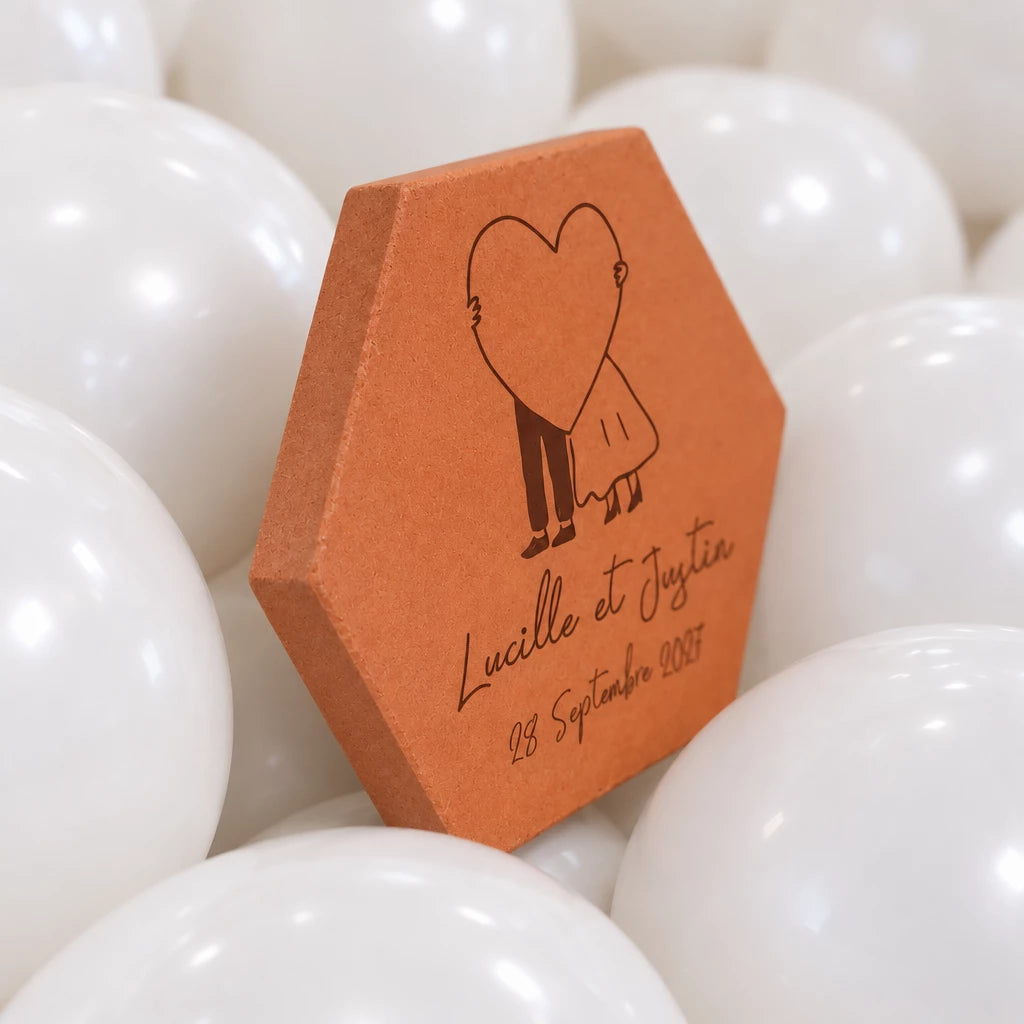 Pieza hexagonal de terracota «Gran corazón» vista de tres cuartos, con el canto visible, entre globos blancos: pareja tras un corazón y «Lucille et Justin».