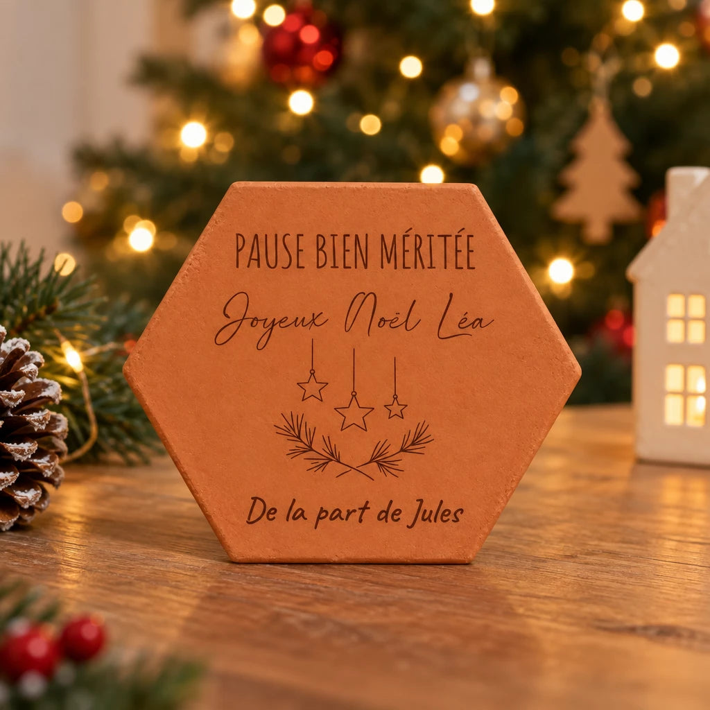 Pieza hexagonal de terracota «Estrellas de Navidad» sobre una mesa delante de un árbol de Navidad iluminado y una casita luminosa.
