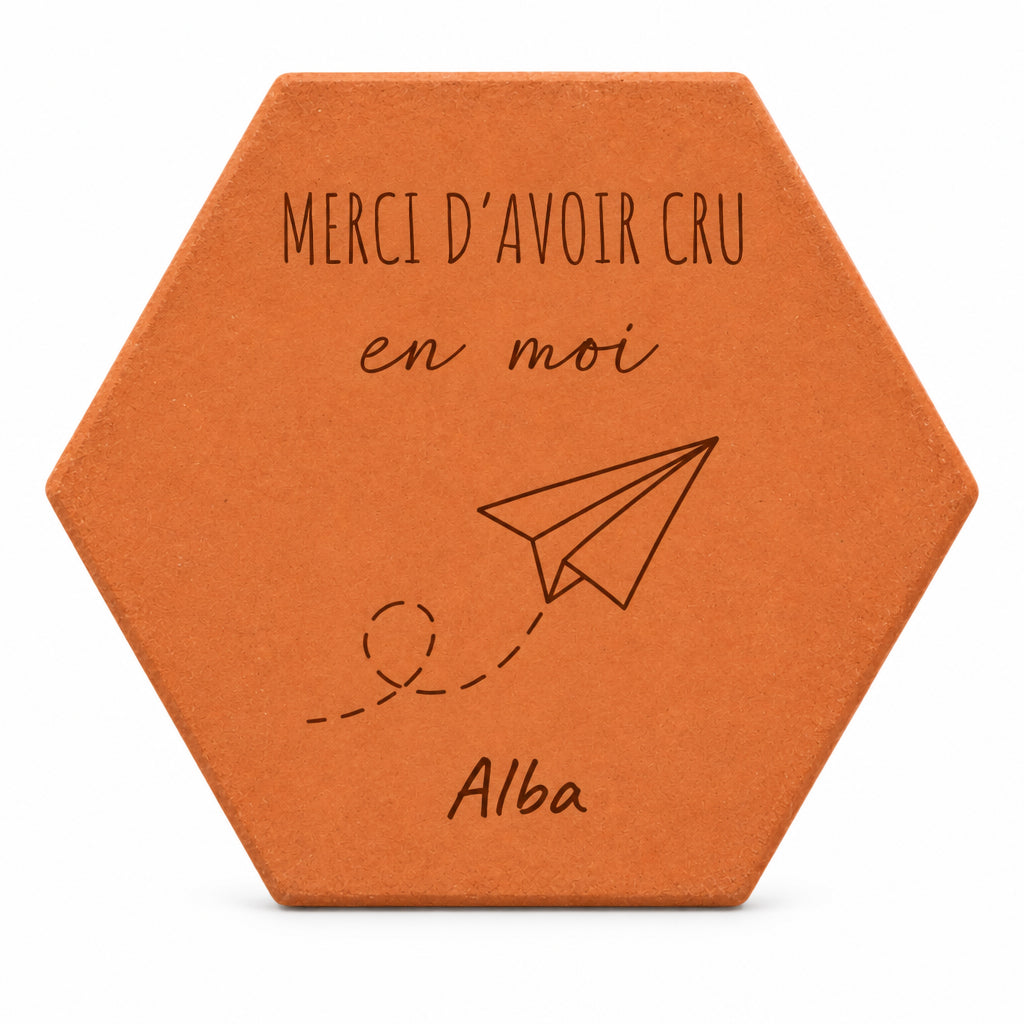 Pieza hexagonal de terracota grabada con «Merci d'avoir cru en moi», un avión de papel con su estela de puntos y el nombre Alba, sobre fondo blanco.